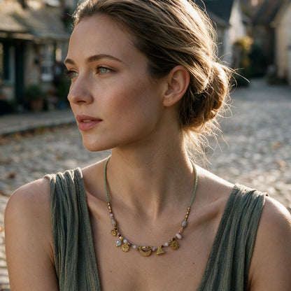 A woman with her hair in a low bun is shown in profile wearing a delicate gold necklace with small charms, including a crescent moon and a small triangle, on a cobblestone street.