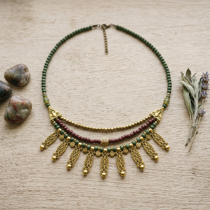 A bohemian-style beaded necklace with green, burgundy, and gold beads, featuring dangling gold filigree leaf-shaped charms, displayed on a light wood background with decorative stones and dried lavender.