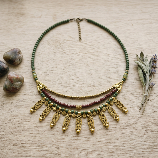 A bohemian-style beaded necklace with green, burgundy, and gold beads, featuring dangling gold filigree leaf-shaped charms, displayed on a light wood background with decorative stones and dried lavender.
