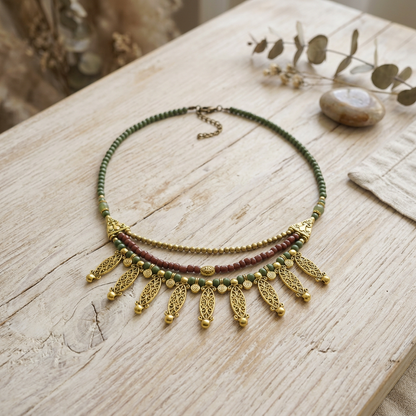A bohemian-style beaded necklace with green and dark red beads, gold-toned filigree leaf pendants, and a matching chain extender, displayed on a rustic light wood surface with dried eucalyptus leaves.
