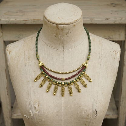 A two-layer statement necklace displayed on a vintage fabric mannequin bust. The necklace features green beads, a central strand of maroon beads, and ornate gold-toned filigree leaf-shaped pendants with small gold bells at the tips.