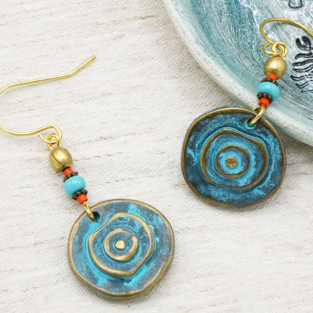 Pair of blue and gold spiral earrings on a textured surface with a ceramic dish in the background.