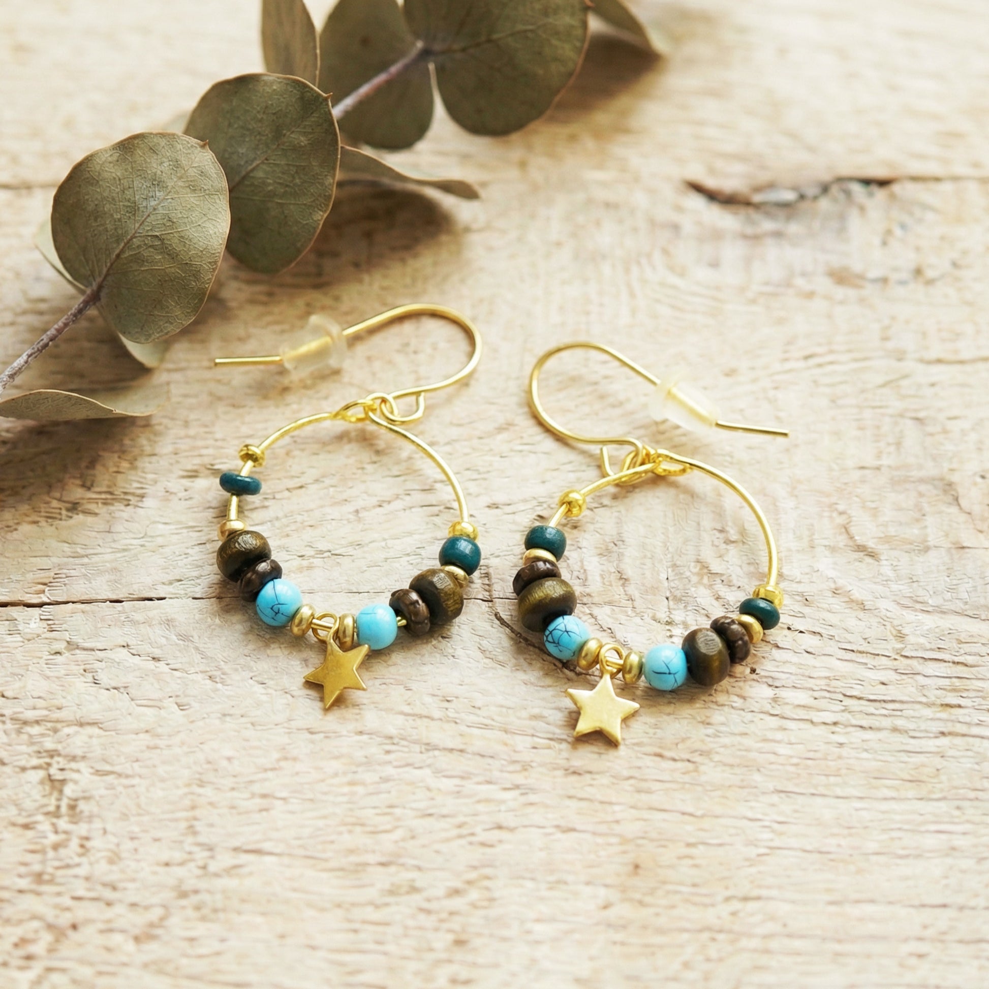 Gold hoop earrings with beads and star charms on a wooden surface with eucalyptus leaves.