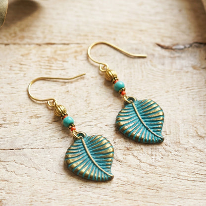 A pair of bohemian-style dangle earrings featuring textured turquoise-blue leaf charms with gold-toned detailing, accented by small turquoise and red beads on gold hooks, displayed on a light wood background.
