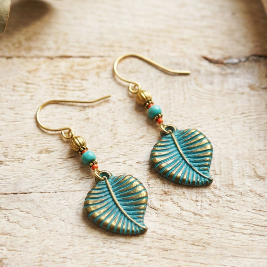 A pair of bohemian-style dangle earrings featuring textured turquoise-blue leaf charms with gold-toned detailing, accented by small turquoise and red beads on gold hooks, displayed on a light wood background.