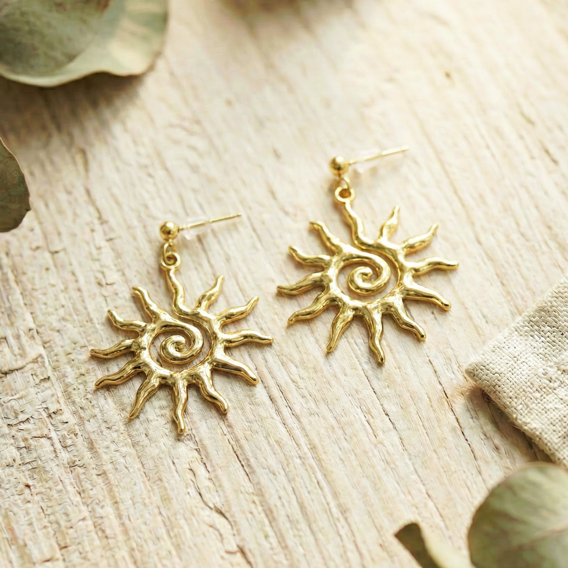 Gold sun-shaped earrings on a wooden surface with leaves.