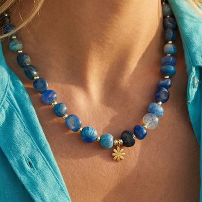 Blue beaded necklace with a gold flower pendant on a person wearing a turquoise top.