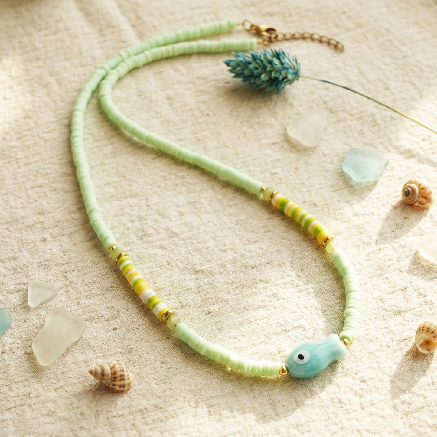Necklace with green and yellow beads on a sandy surface with shells and sea glass.