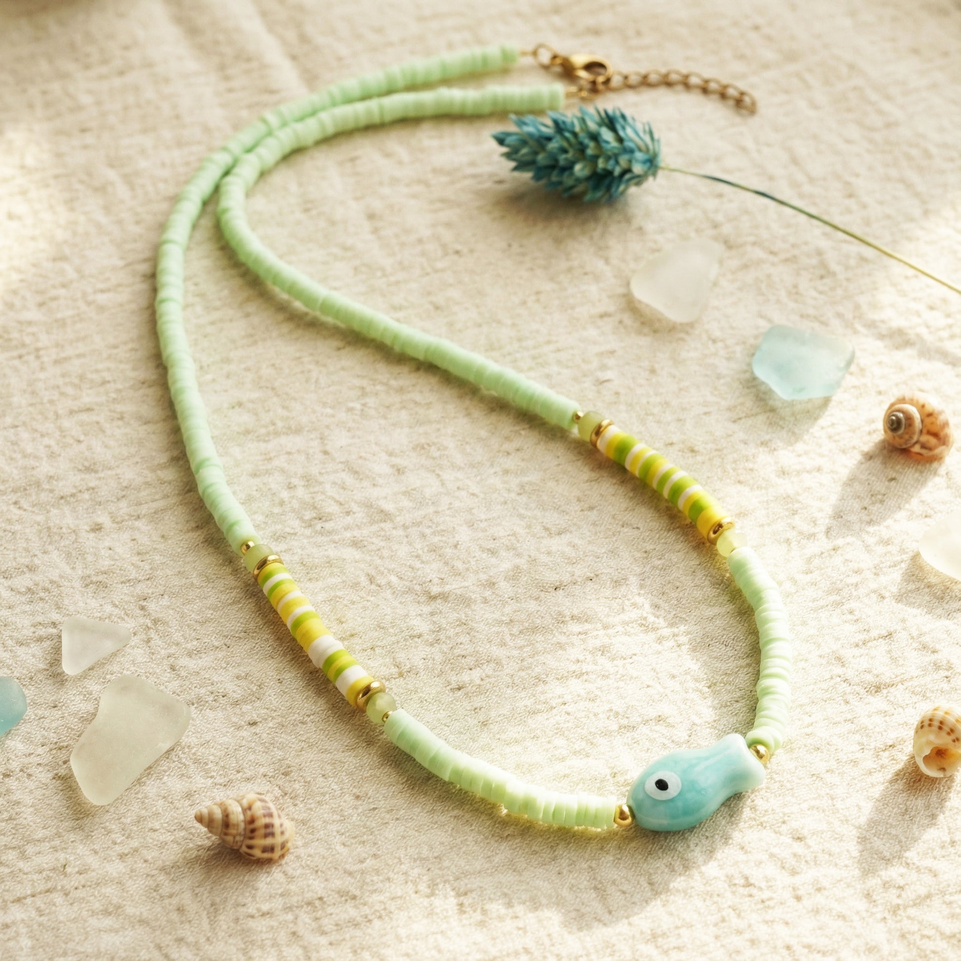 Necklace with green and yellow beads on a sandy surface with shells and sea glass.