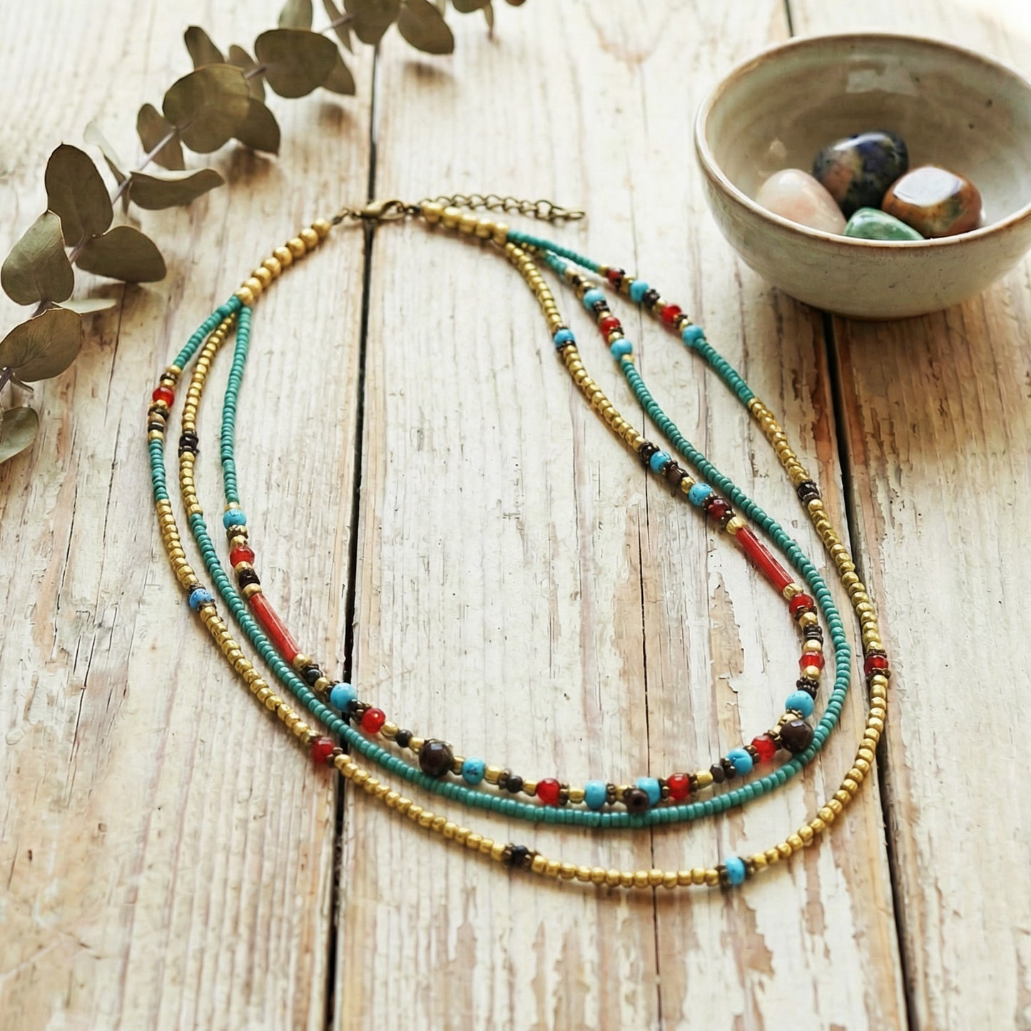 Colorful beaded necklace on a wooden surface with a bowl of stones and leaves.