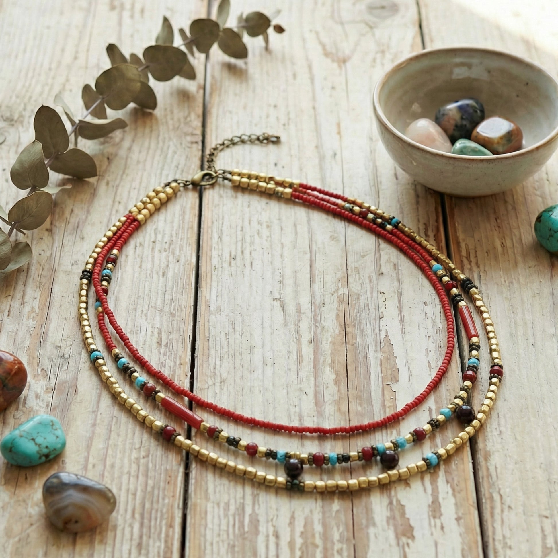 Multistrand beaded necklace on a wooden surface with a bowl of stones and eucalyptus leaves.