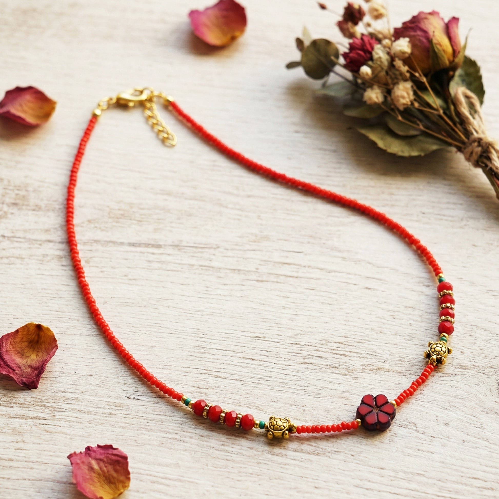 Red beaded necklace with floral elements on a light wooden surface with dried flowers.