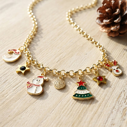 A gold-tone holiday charm necklace displayed on a light wood surface, featuring snowman, Christmas tree, star, and snowman charms, with a pinecone prop in the corner.