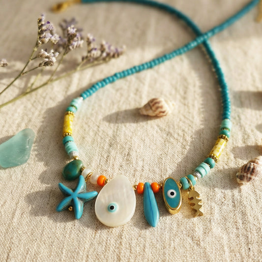 Necklace with blue and gold beads and charms on a textured surface with dried flowers and shells.