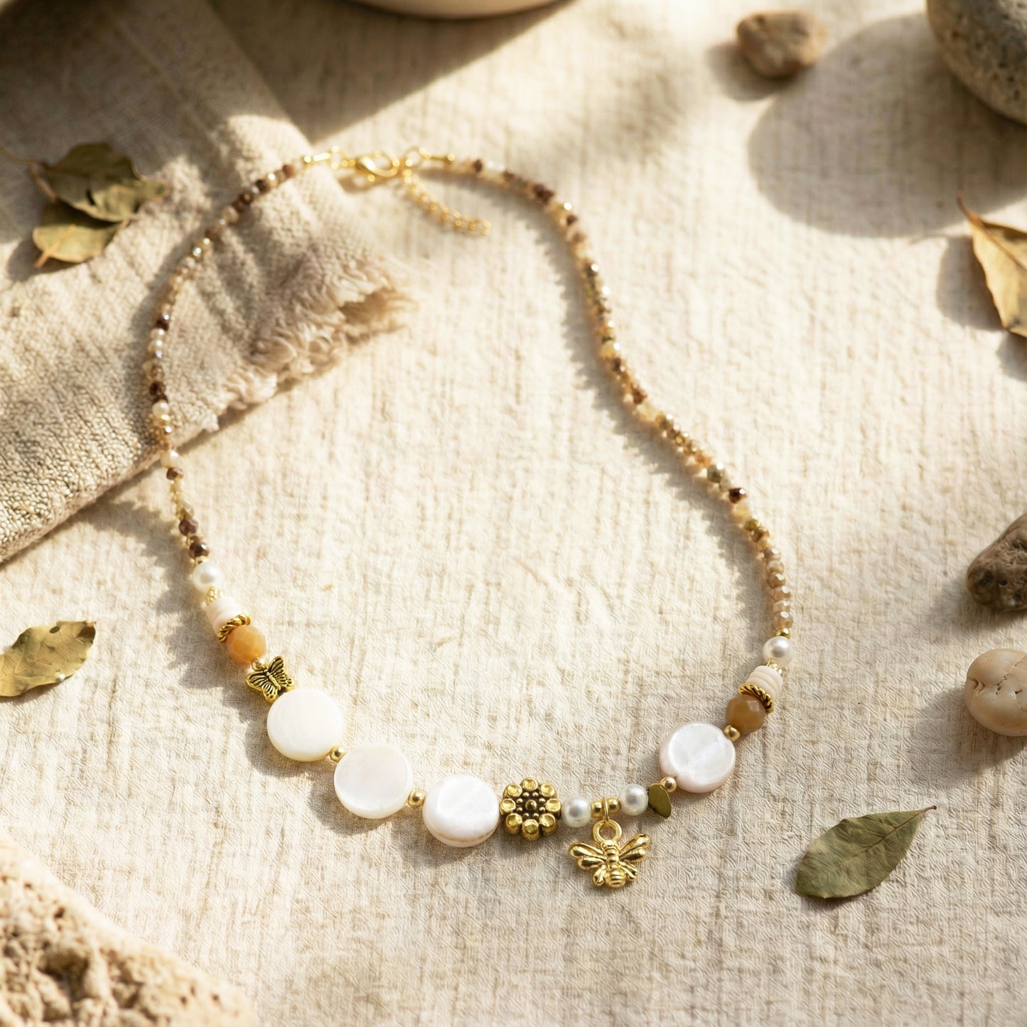 Necklace with white beads and gold charms on a textured beige surface with leaves and stones.