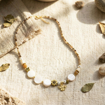 Necklace with white beads and gold charms on a textured beige surface with leaves and stones.