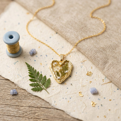 A gold heart-shaped pendant necklace with dried green leaves and small white pearls inside, displayed on a textured beige fabric with a spool of gold thread, a green fern leaf, and small blue stones.