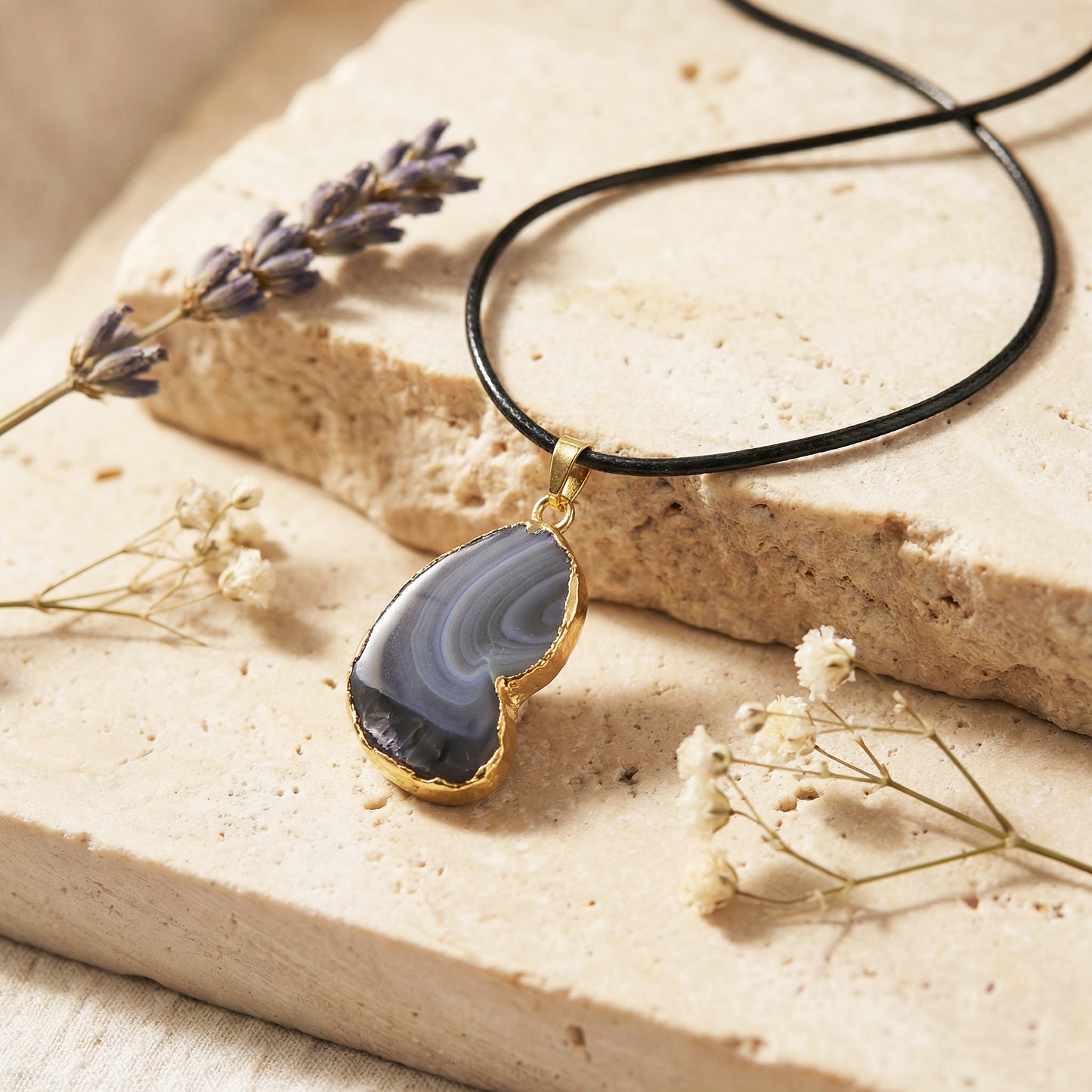 Necklace with a blue agate pendant on a beige stone surface with dried flowers.