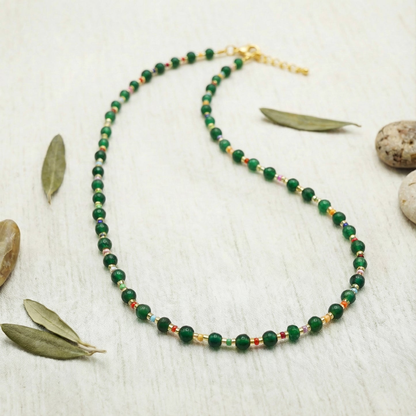 Green beaded necklace on a light wooden surface with leaves and stones.