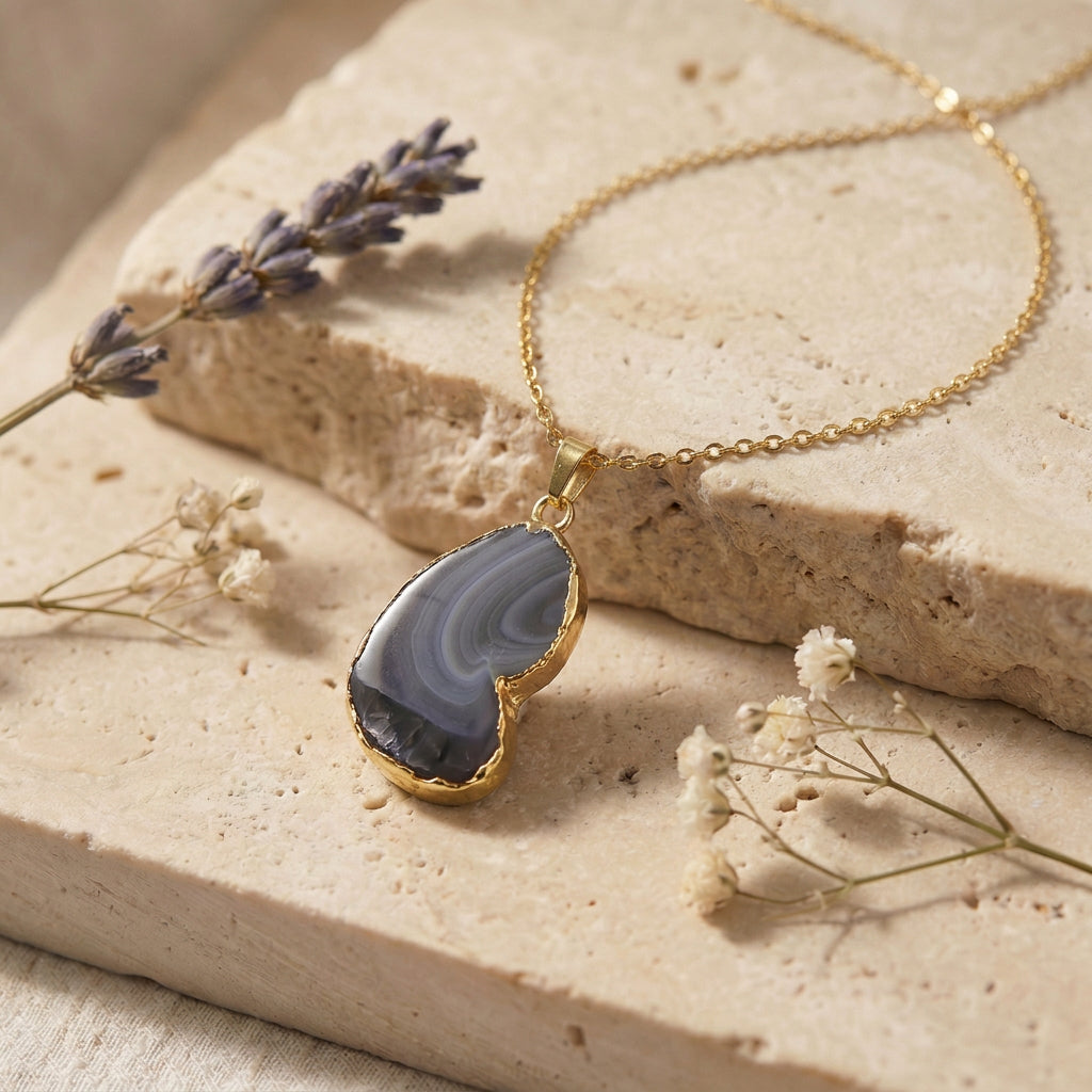 A gold necklace with a polished agate stone pendant, styled on a textured beige stone slab with dried lavender and baby's breath flowers.