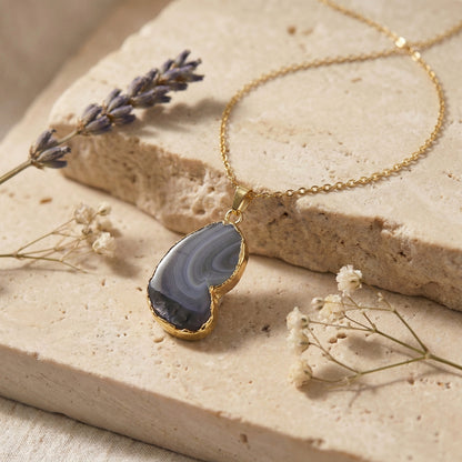 A gold necklace with a polished agate stone pendant, styled on a textured beige stone slab with dried lavender and baby's breath flowers.