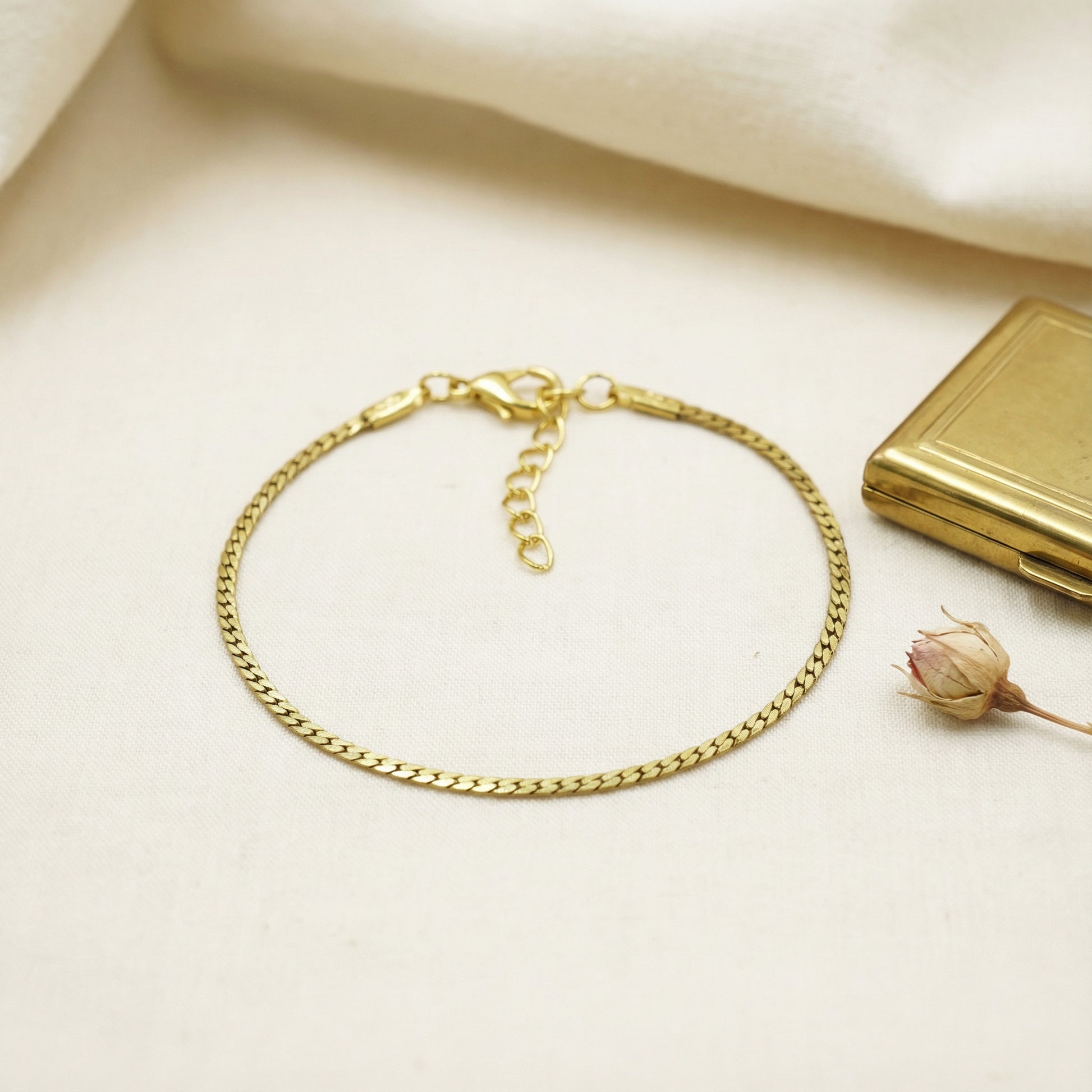 Gold chain bracelet on a light beige surface with a small gold box and dried flower.