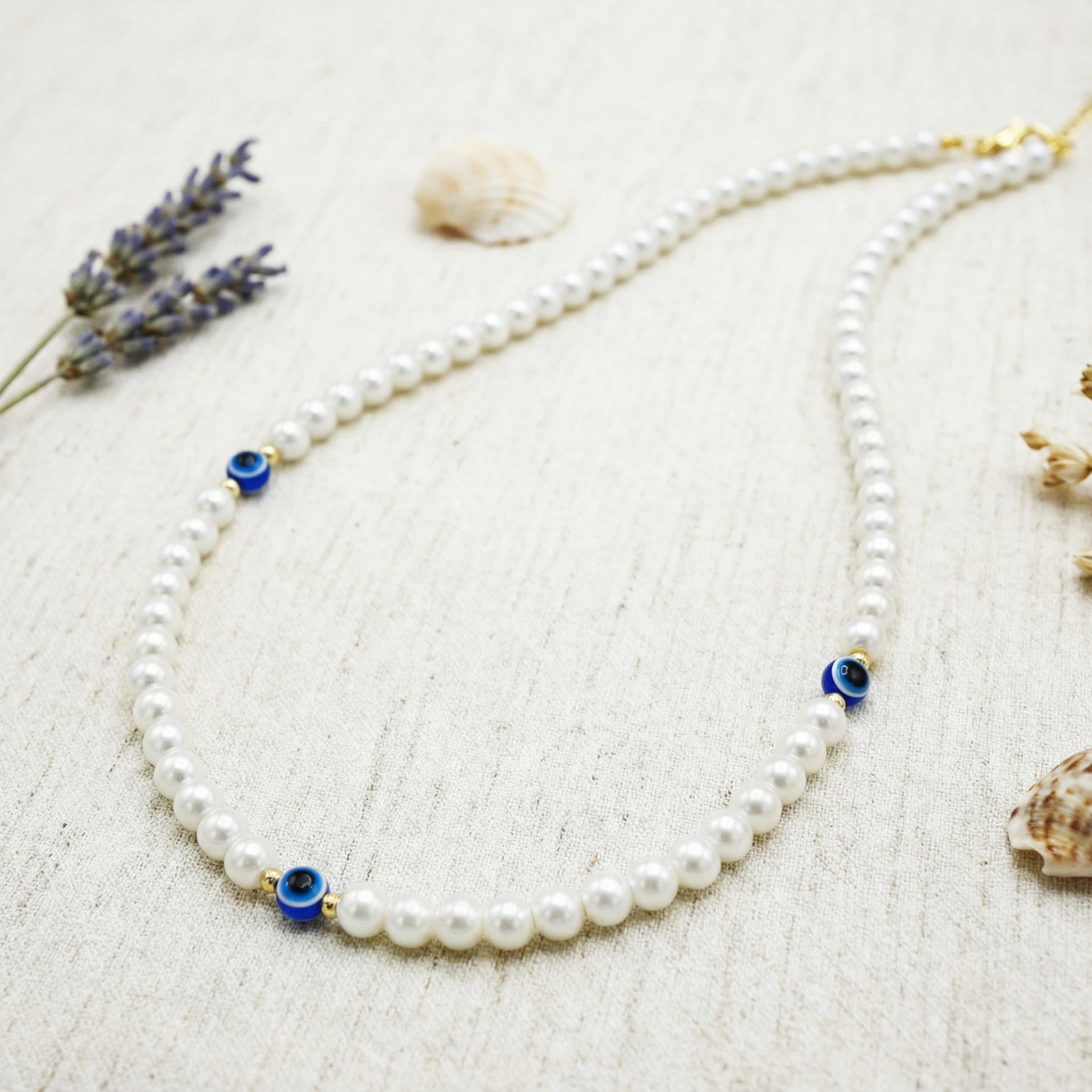 Pearl necklace with blue evil eye beads on a textured surface with lavender and shells.