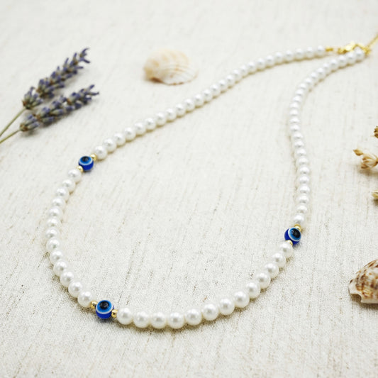 Pearl necklace with blue evil eye beads on a textured surface with lavender and shells.