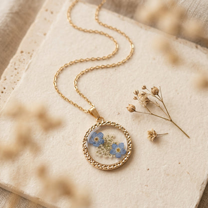 A gold chain necklace with a circular pendant featuring small blue and white dried flowers, styled on a textured cream stone slab with blurred dried flowers.