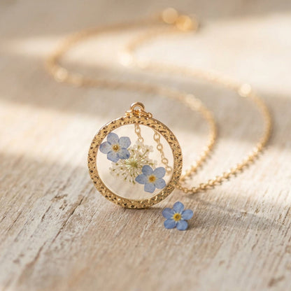 A gold-tone round pendant necklace with dried blue and white flowers under clear cover, displayed on a light wood surface with a matching flower charm nearby.