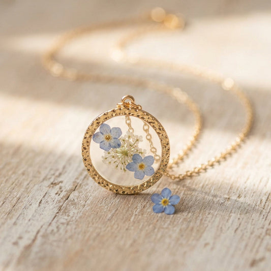 A gold-tone round pendant necklace with dried blue and white flowers under clear cover, displayed on a light wood surface with a matching flower charm nearby.