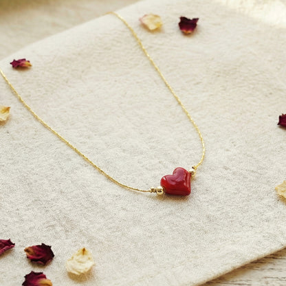 Gold necklace with a red heart pendant on a textured surface with dried flowers.