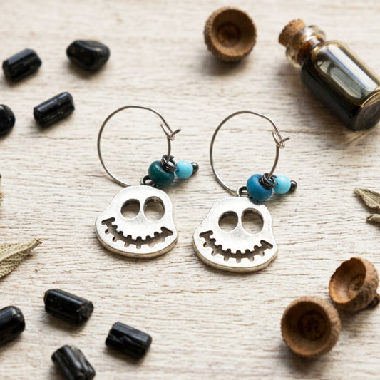 A pair of silver ghost hoop earrings with small turquoise blue beads, displayed on a light wood surface with Halloween-themed props like black stones and dried leaves.