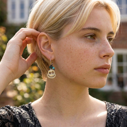 A close-up lifestyle shot of a woman with blonde hair wearing a dangling ghost earring. The earring features a gold skull charm with blue and green beads above it.
