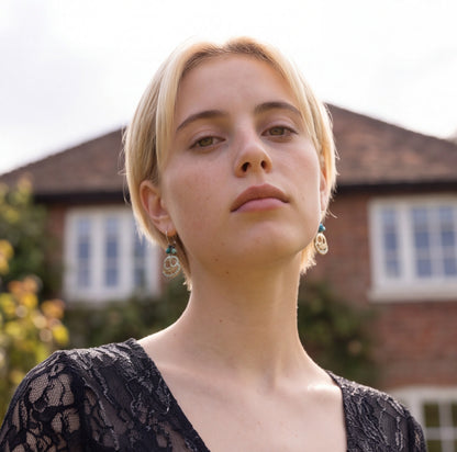 A close-up portrait of a woman with short blonde hair wearing dangling ghost earrings and a black lace top, photographed outdoors with a blurred brick house in the background.