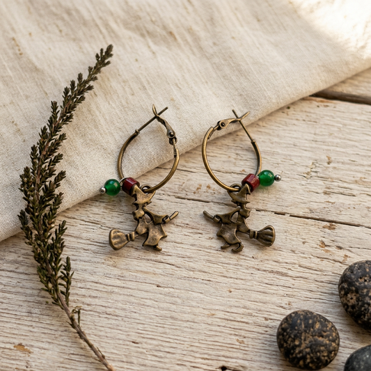 A pair of antique bronze hoop earrings with witch charms and green beads, displayed on a rustic wooden surface with a linen cloth and dried botanicals.