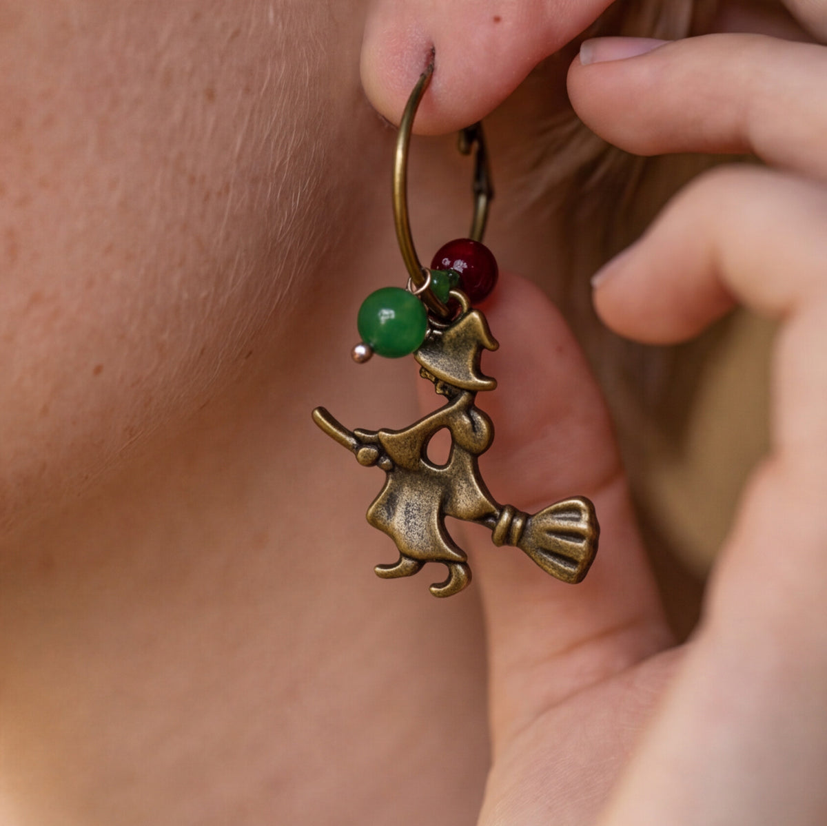 A close-up of a person wearing a dangling earring with an antique bronze witch riding a broomstick charm, accented by a green bead and a red bead.