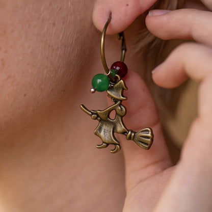 A close-up of a person wearing a dangling earring with an antique bronze witch riding a broomstick charm, accented by a green bead and a red bead.