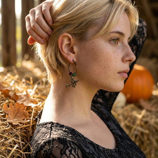 A woman in profile wearing a black lace top and a dangling gothic-style earring with a witch charm, set against a rustic autumn background of straw, leaves, and a pumpkin.