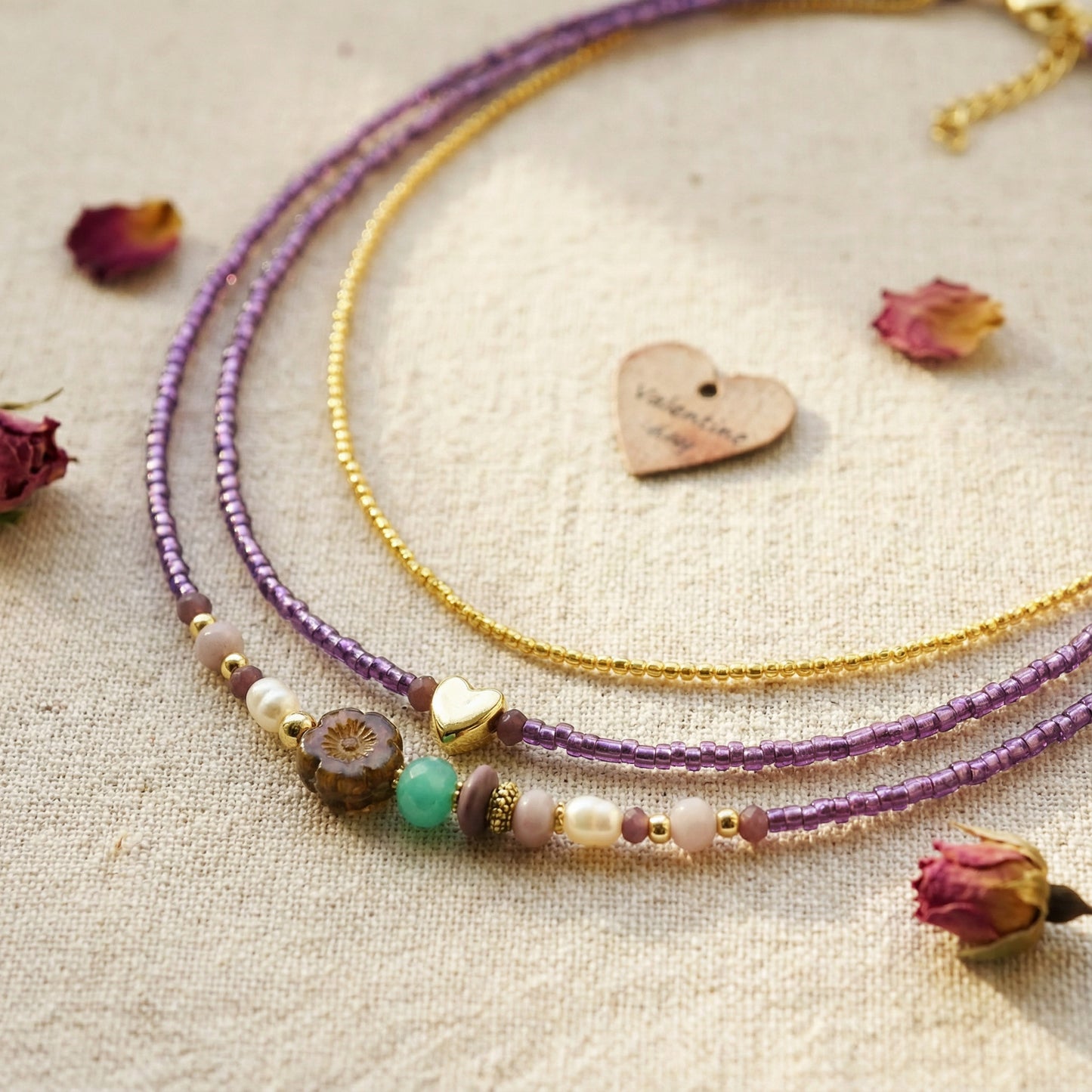 Two beaded necklaces, one purple and one gold, on a textured surface with dried flowers.