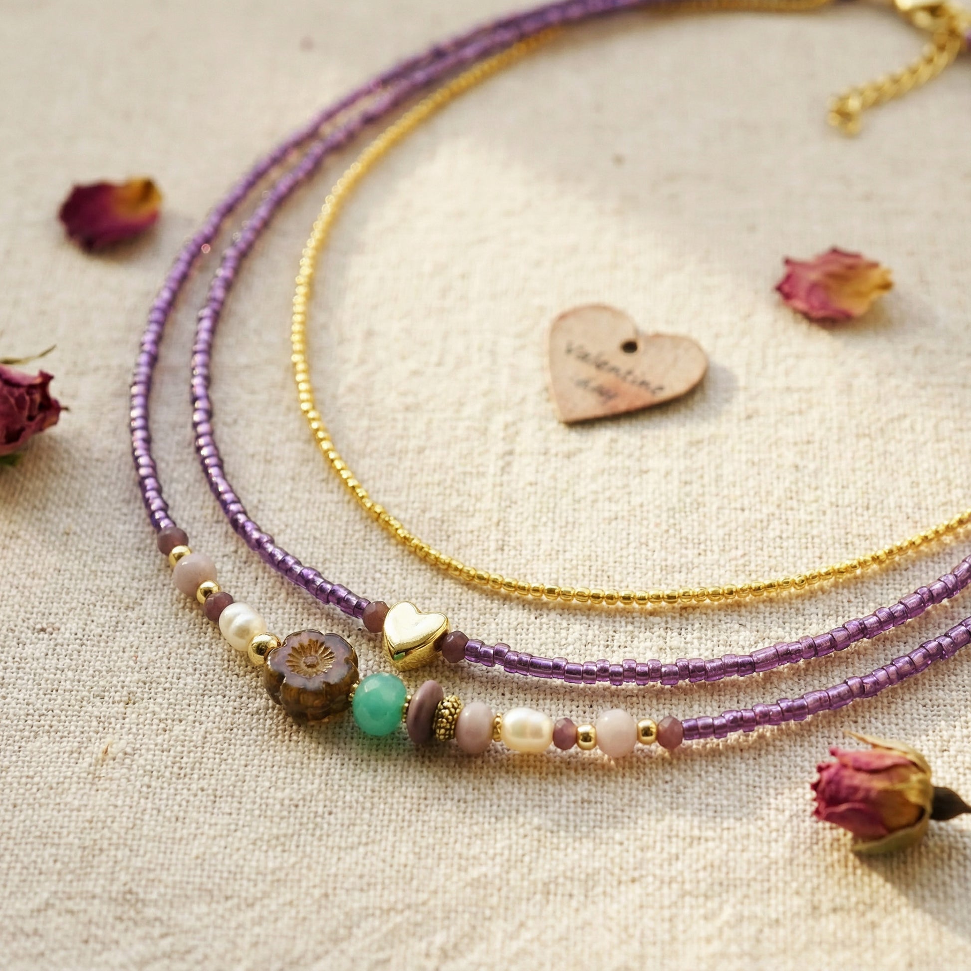 Two beaded necklaces, one purple and one gold, on a textured surface with dried flowers.