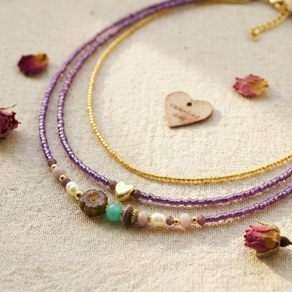 Two beaded necklaces, one purple and one gold, on a textured surface with dried flowers.