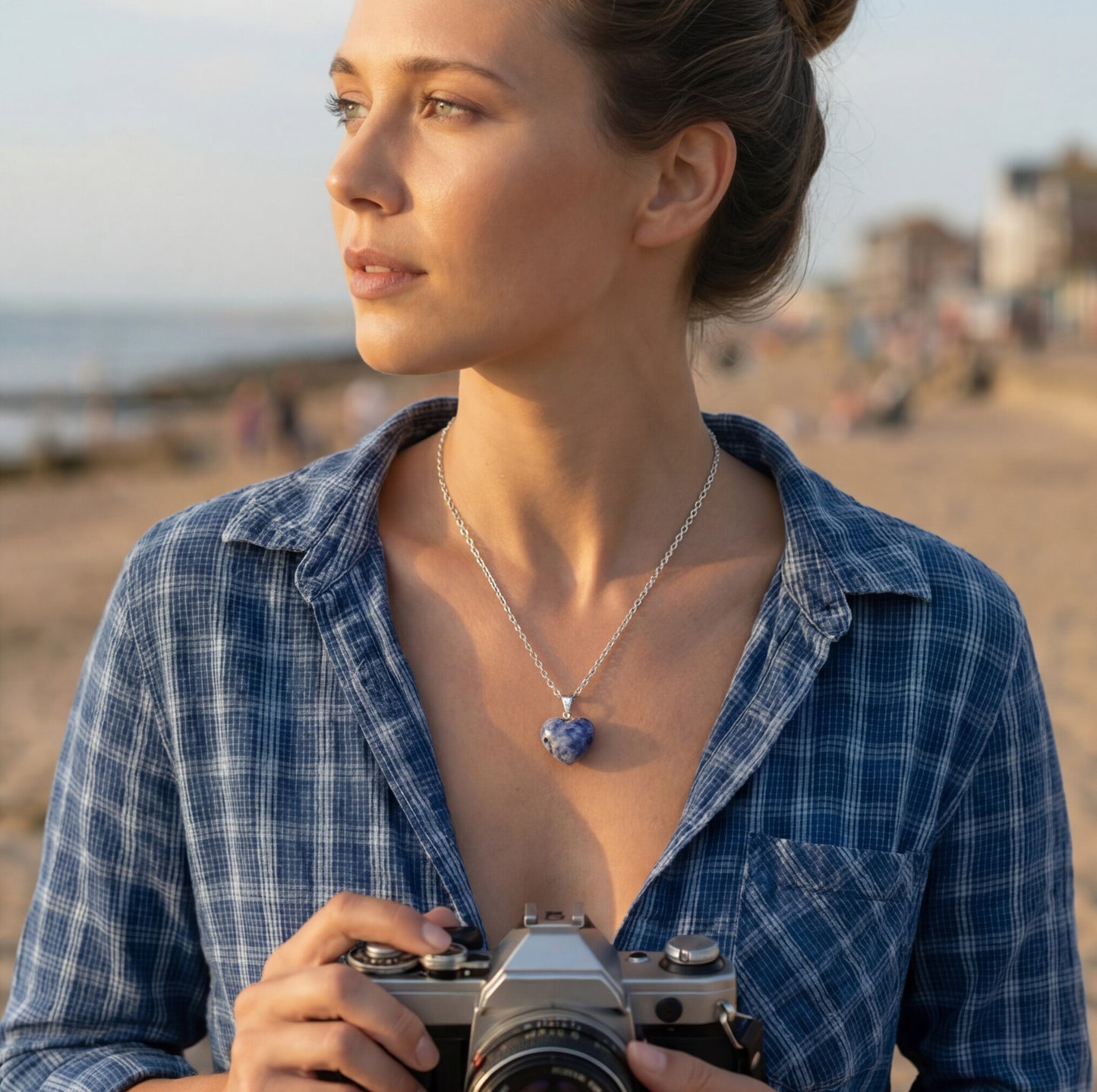 Woman holding a camera on a beach with a necklace featuring a blue pendant.