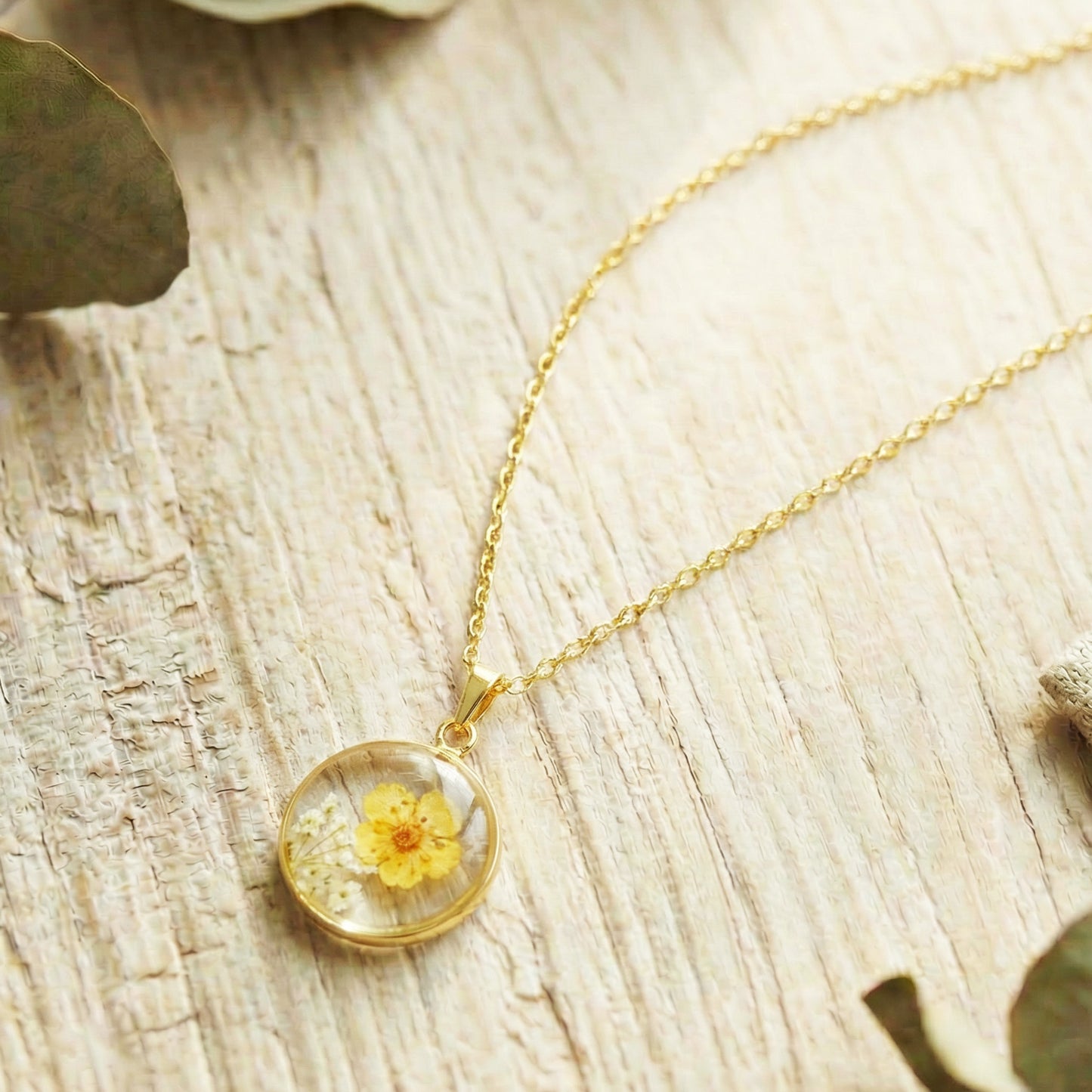Gold necklace with a clear pendant featuring a yellow flower on a wooden surface.