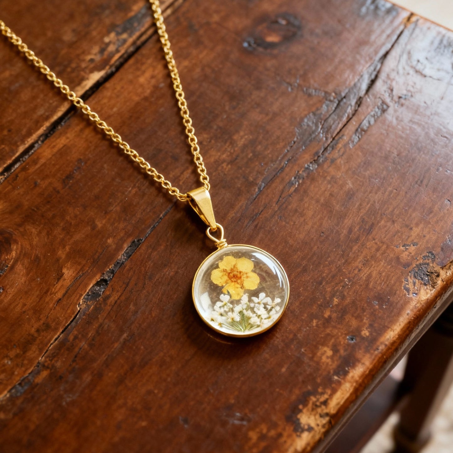 Gold necklace with a round pendant featuring a yellow flower and white blossoms on a wooden surface.