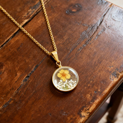 Gold necklace with a round pendant featuring a yellow flower and white blossoms on a wooden surface.
