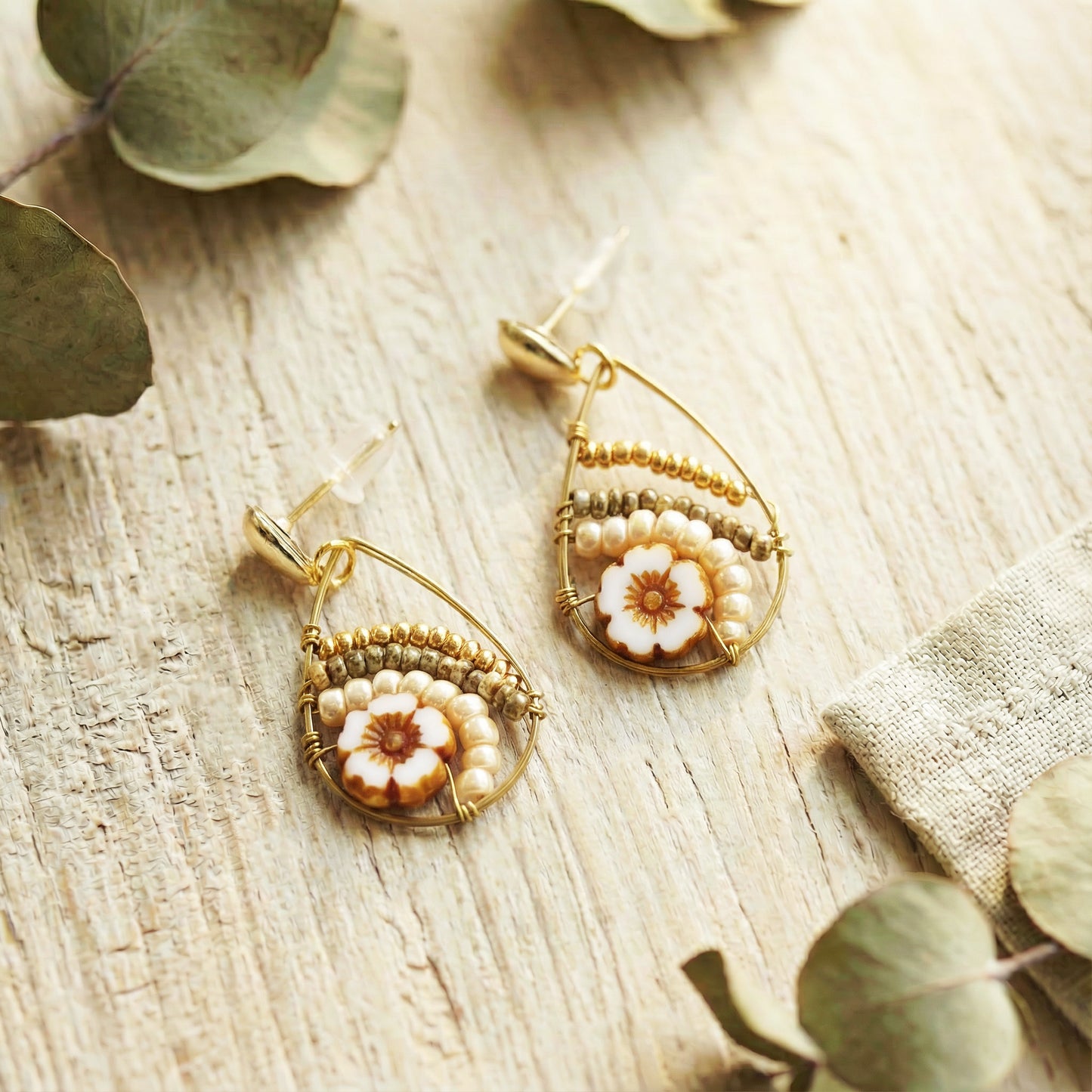 Gold earrings with floral designs on a wooden surface with eucalyptus leaves.