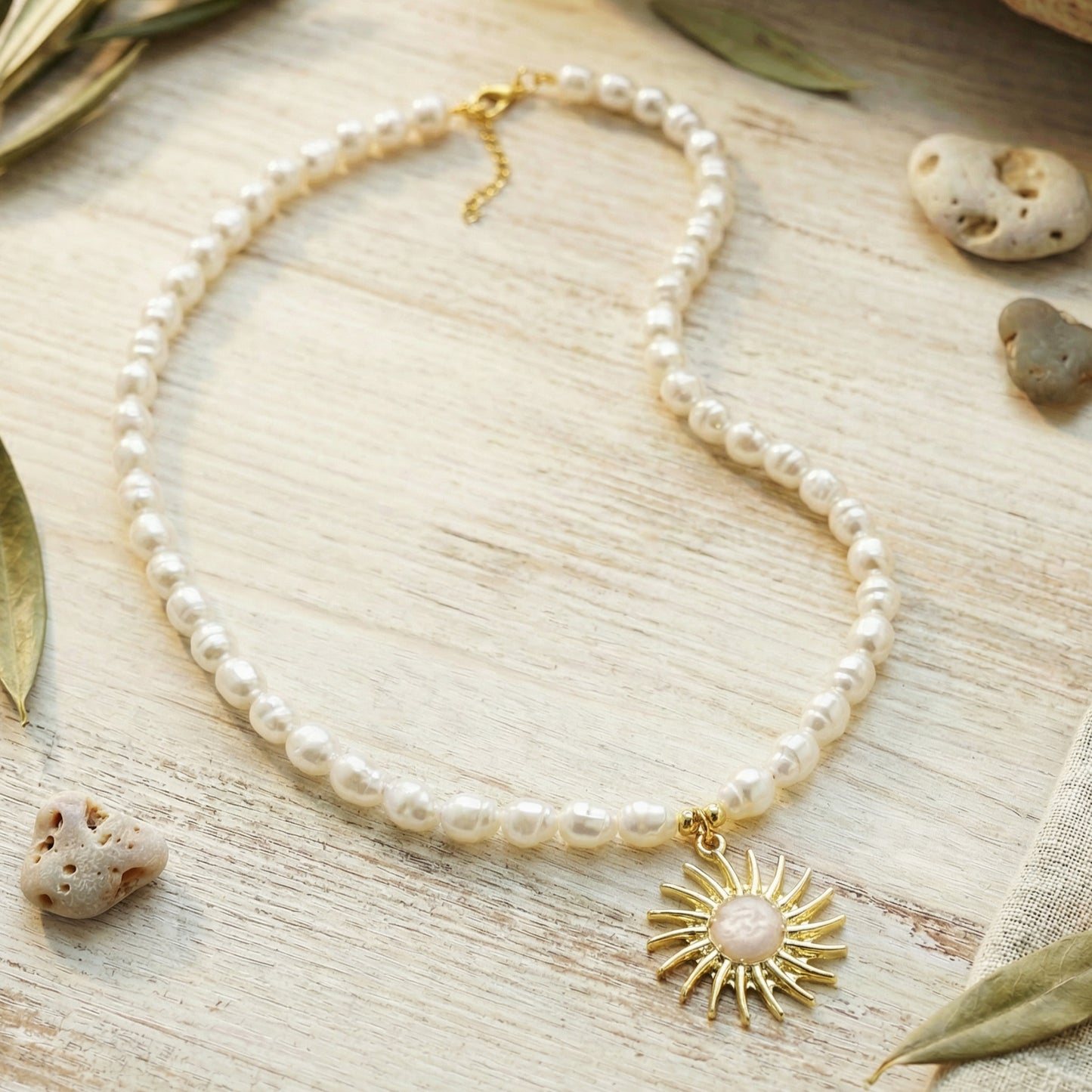 Pearl necklace with a gold sun pendant on a natural wooden surface with rocks and leaves.