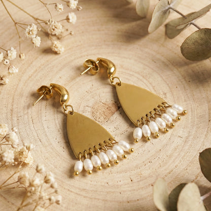 Gold earrings with pearl drops on a wooden surface with dried flowers and leaves.