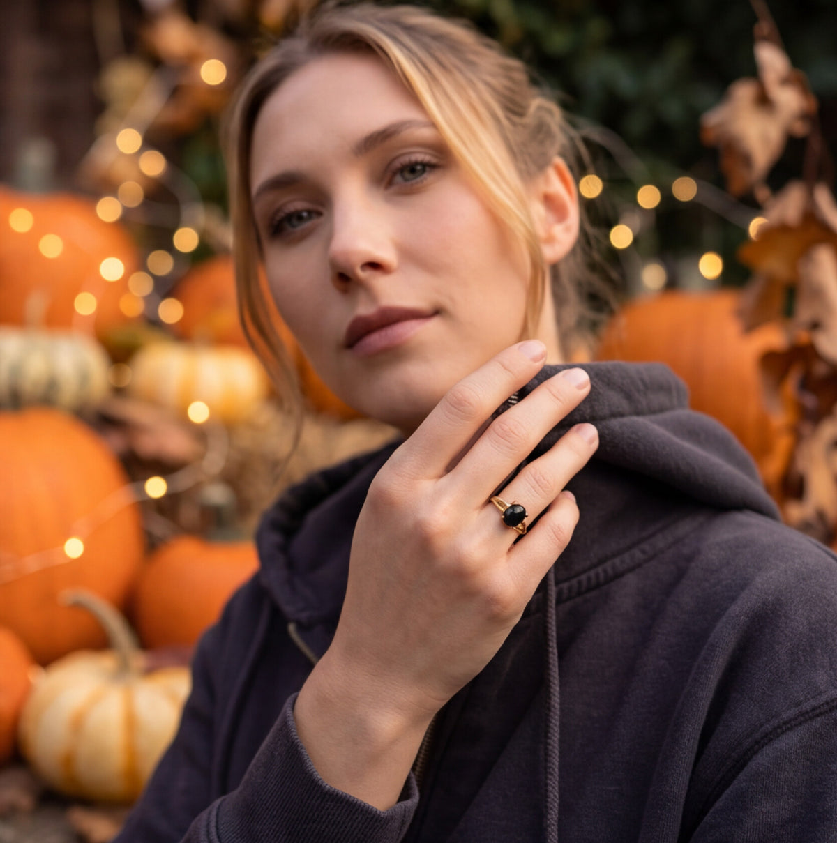 A woman wearing a black onyx ring with a dark stone, posing in an autumn setting with pumpkins and warm string lights.